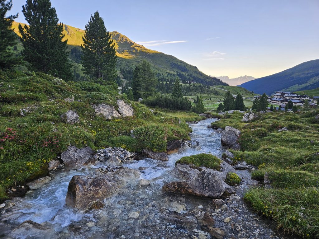Bachlauf mit Felsen, Blumen und Hütten im Hintergrund bei Sonnenaufgang