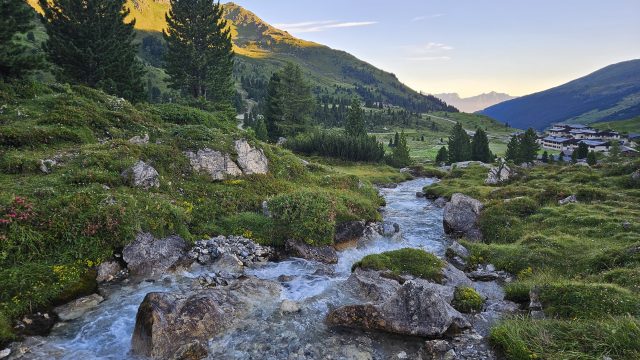 Bachlauf mit Felsen, Blumen und Hütten im Hintergrund bei Sonnenaufgang