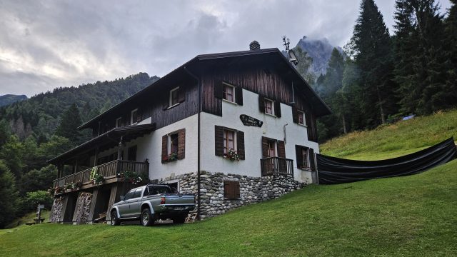 Rifugio Bianchet vor bewaldetem Berghang bei Dämmerung, traditionelle Berghütte mit Holzverkleidung und Steinmauer